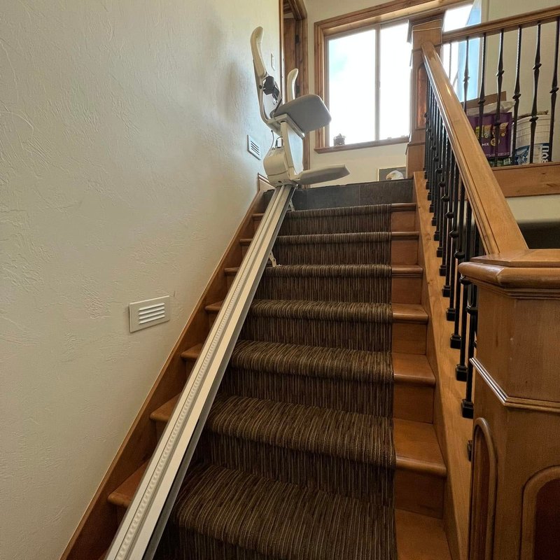 Stair lift rail running up a long carpeted staircase to the lift parked at the top