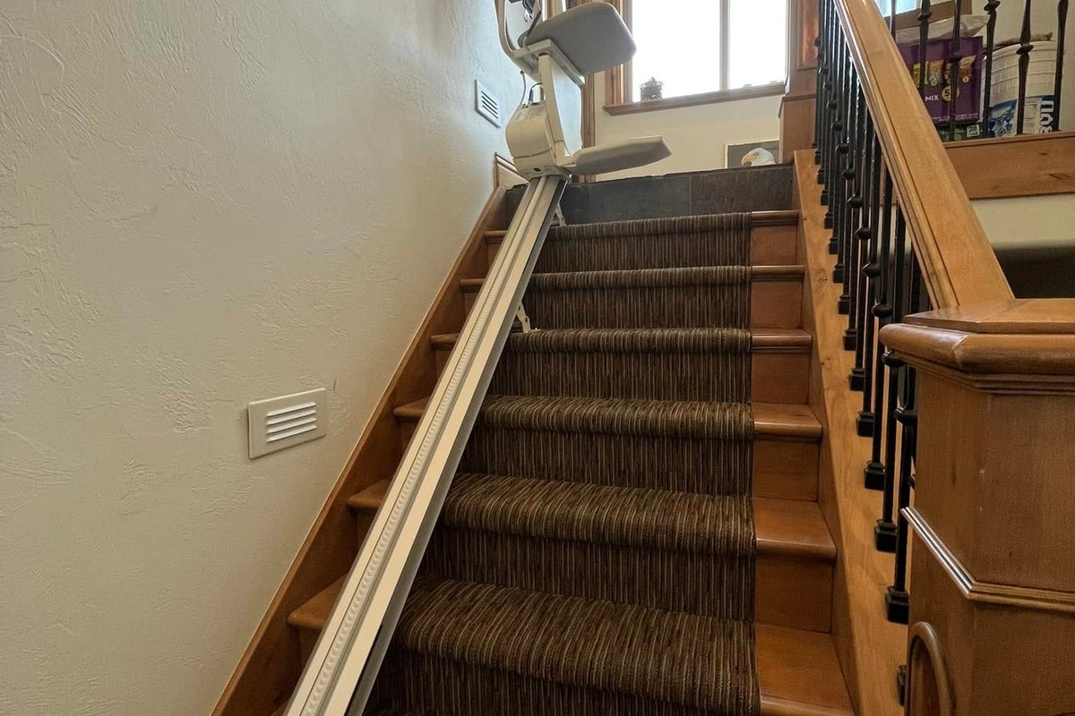 Stair lift parked at the top landing of a long carpeted staircase with its rail running up the wall