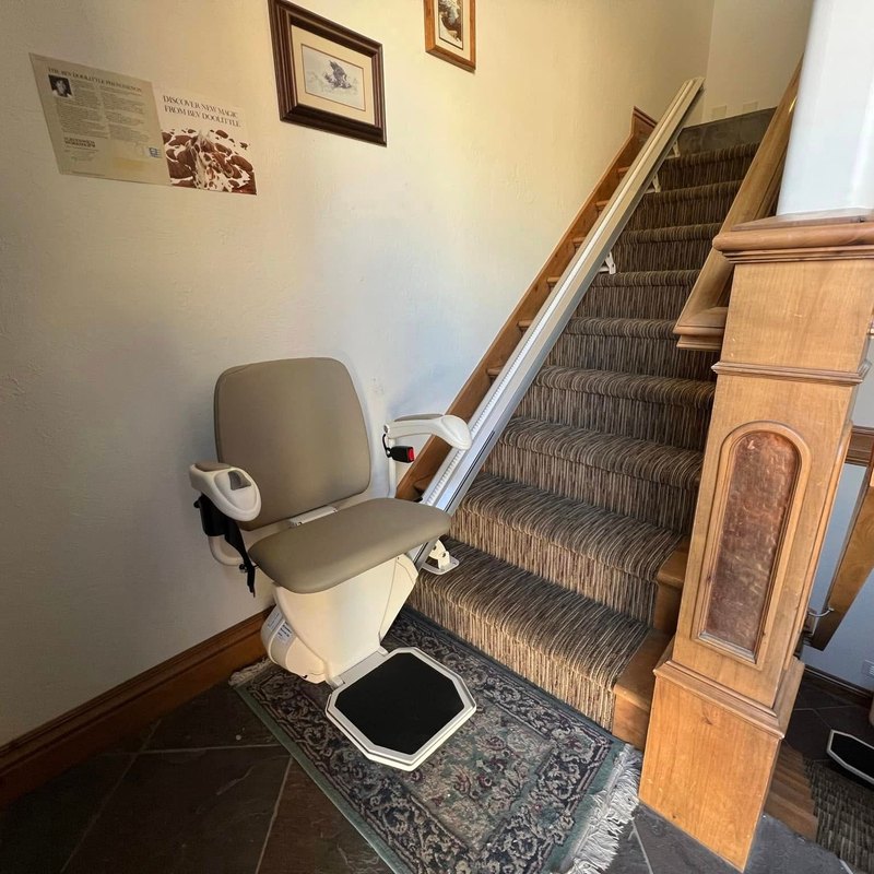 Stair lift at the bottom of a carpeted staircase beside a carved wood newel post